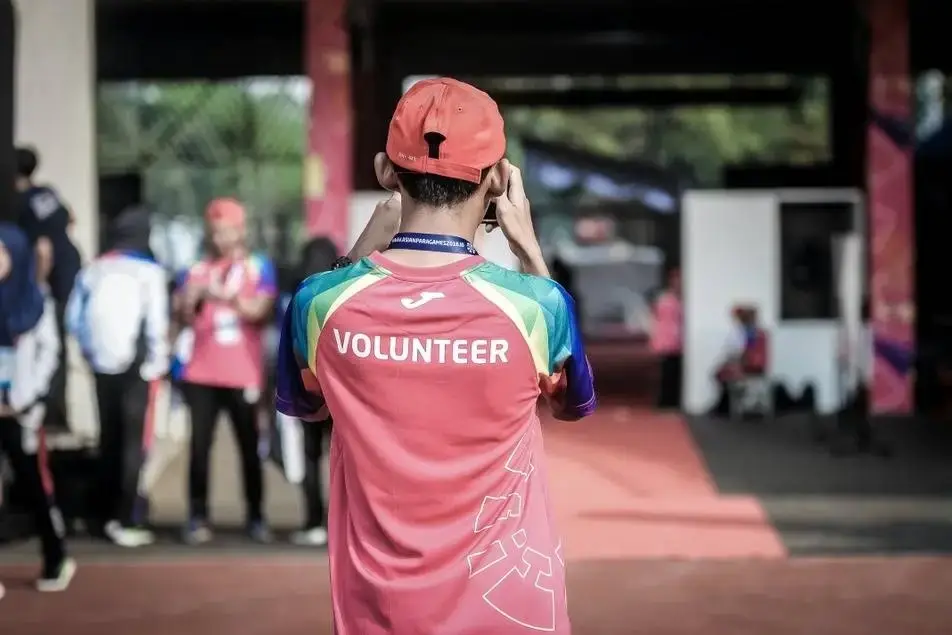 Legenda: Voluntário de costas, trajando boné e blusa vermelha, tirando foto do ambiente. Ele está localizado em um local aberto, aparentemente esportivo, com outros voluntários ao fundo.