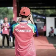 Legenda: Voluntário de costas, trajando boné e blusa vermelha, tirando foto do ambiente. Ele está localizado em um local aberto, aparentemente esportivo, com outros voluntários ao fundo.