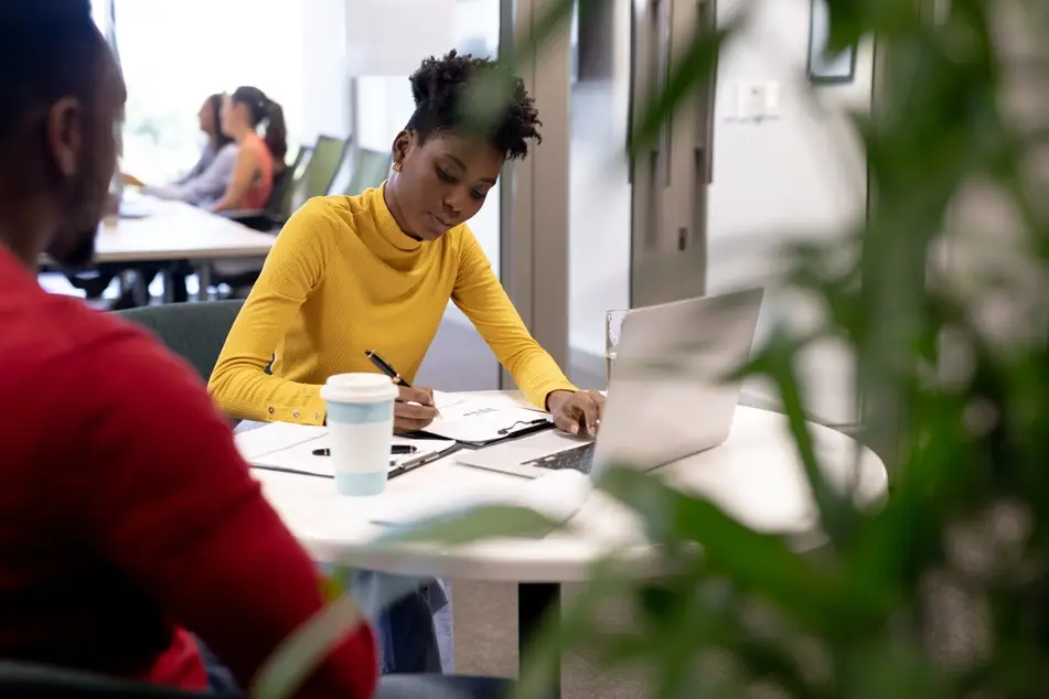 A Black student wearing a bright yellow turtle neck studies at a round table in the library.